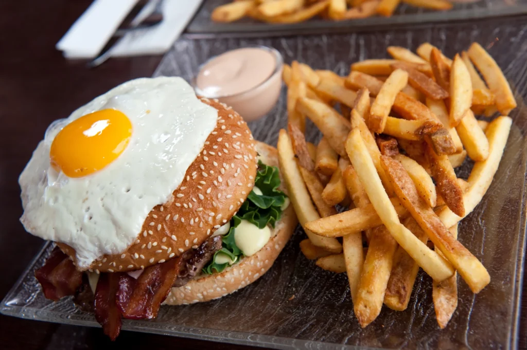 Een grote hamburger met spek, sla en een spiegelei ligt naast een portie friet en een schaaltje saus
