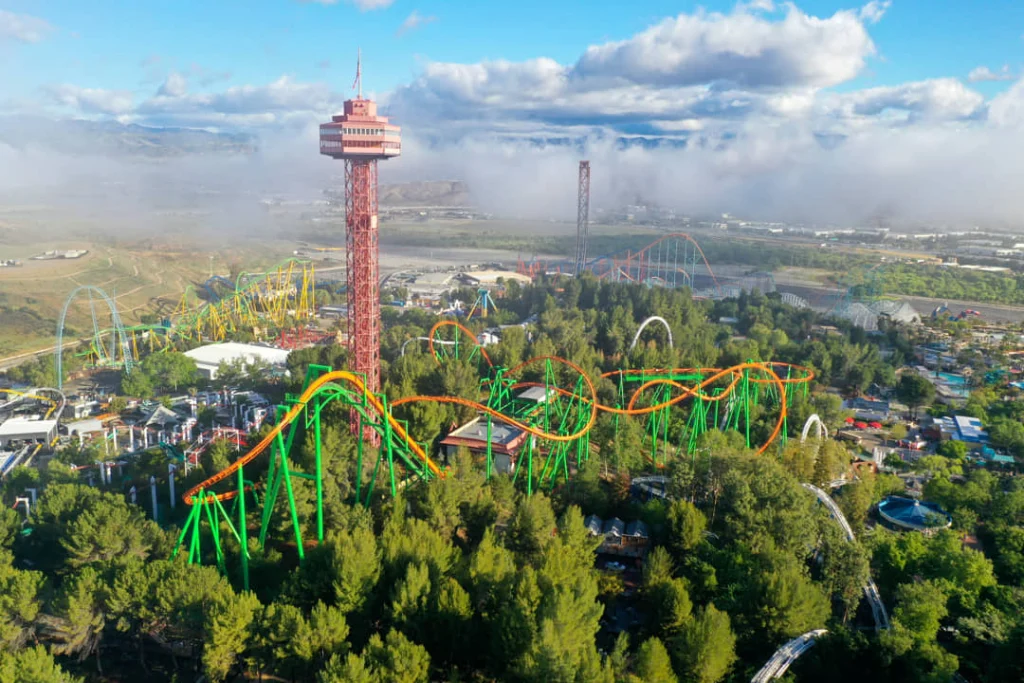 Luchtfoto van een pretpark met een rood witte uitkijktoren tussen kleurrijke achtbanen en bomen, met lage mist op de achtergrond