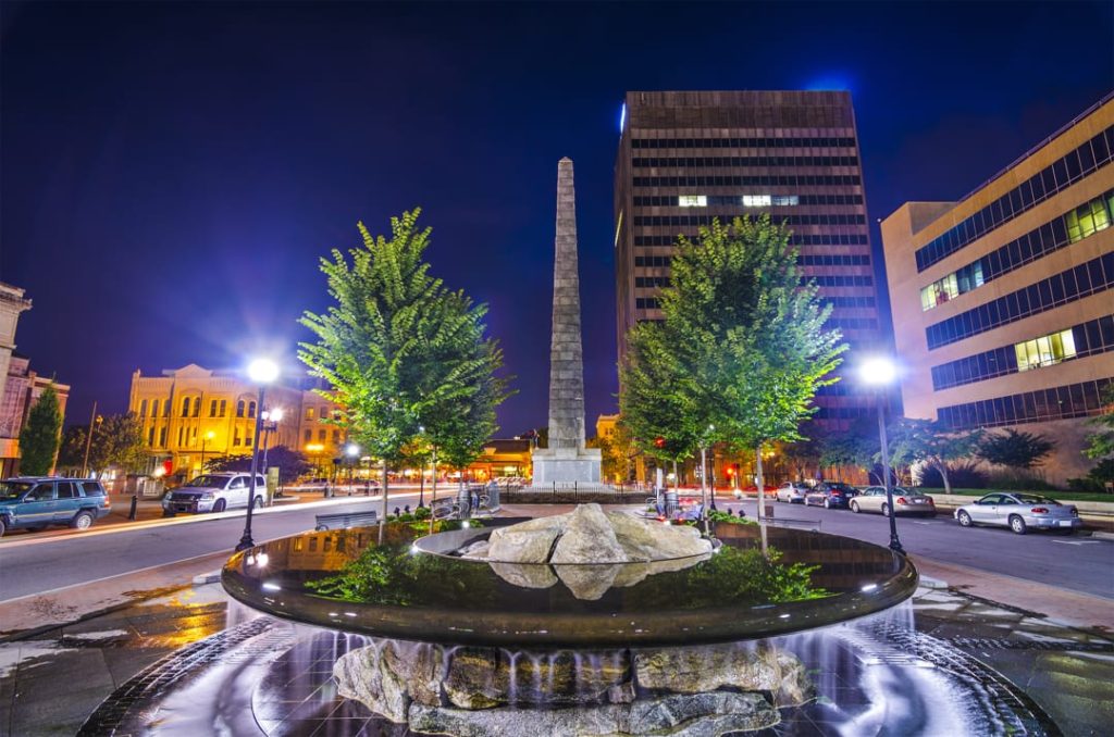 Verlicht plein met een hoge stenen obelisk en een fontein omgeven door bomen en kantoorgebouwen bij nacht