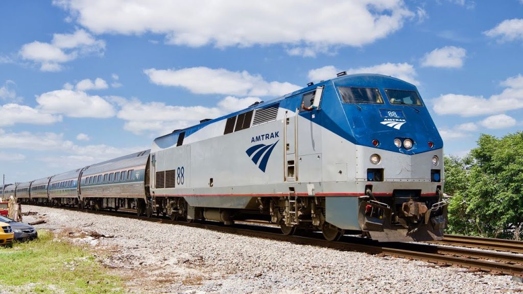 Blauw witte Amtrak locomotief met passagierswagons rijdt over een spoor onder een blauwe lucht met wolken