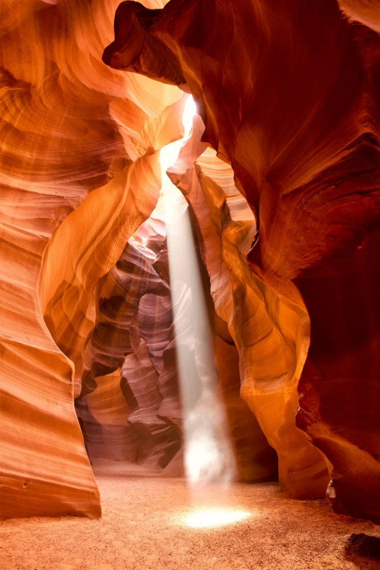 Smalle lichtstraal valt door een opening in de gegolfde oranje rotswanden van Antelope Canyon op het zanderige pad