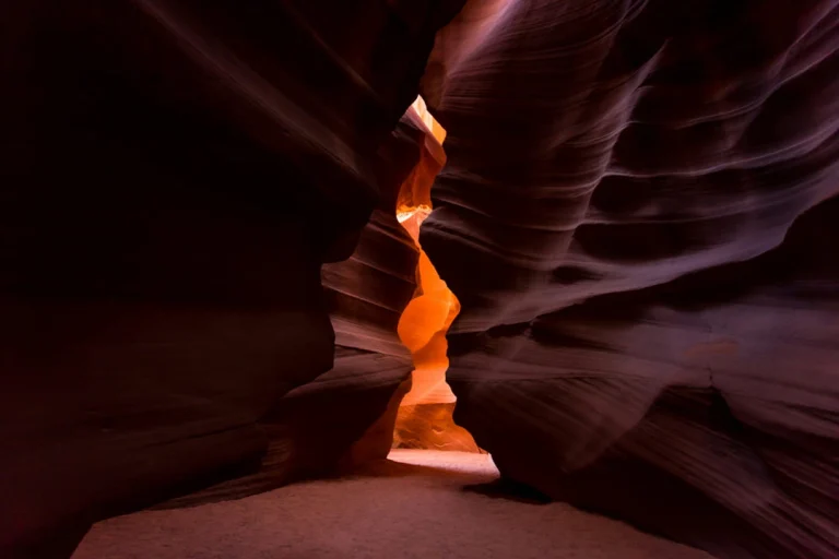 Nauwe doorgang in Antelope Canyon waar warm licht op de golvende oranje rotswanden valt en een bezoeker in de verte zichtbaar is