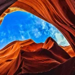 Blik omhoog tussen de golvende roodoranje rotswanden van Antelope Canyon naar een stukje blauwe lucht met wolken