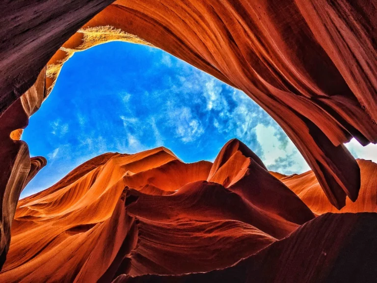 Blik omhoog tussen de golvende roodoranje rotswanden van Antelope Canyon naar een stukje blauwe lucht met wolken