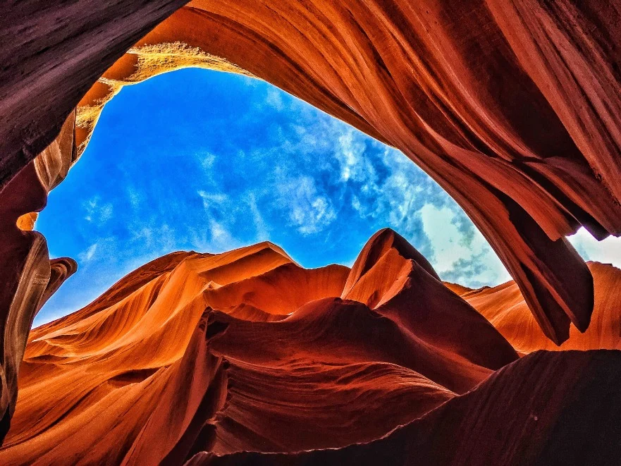 Blik omhoog tussen de golvende roodoranje rotswanden van Antelope Canyon naar een stukje blauwe lucht met wolken
