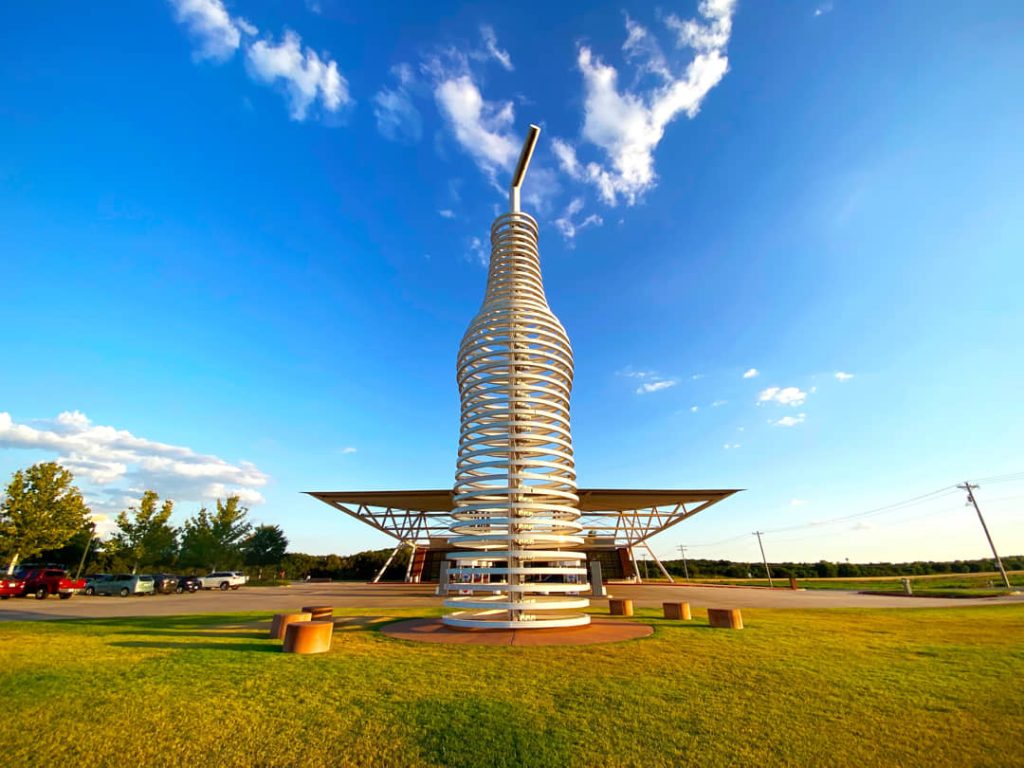 Enorme sculptuur van een glazen frisdrankfles gemaakt van gebogen metalen ringen naast een wegrestaurant onder een blauwe lucht