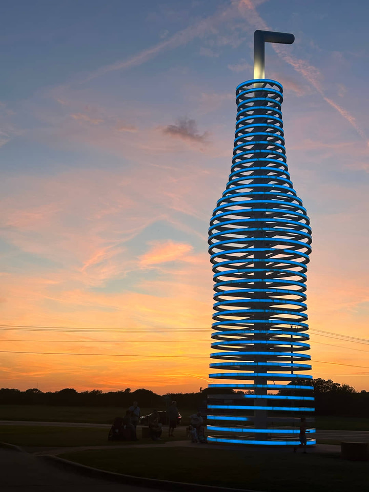 Blauw verlichte sculptuur van een frisdrankfles van metalen ringen tegen een oranje avondlucht met mensen ervoor