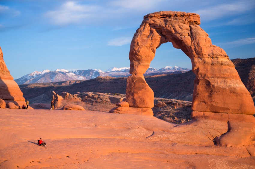 De iconische vrijstaande rotsboog Delicate Arch steekt af tegen besneeuwde bergen met wandelaars op de rotsen eronder in Arches National Park