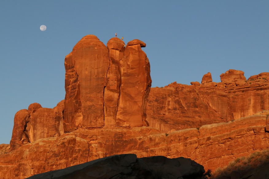 Hoge oranjerode rotstorens steken af tegen een blauwe lucht met de volle maan zichtbaar boven de formatie in Arches National Park