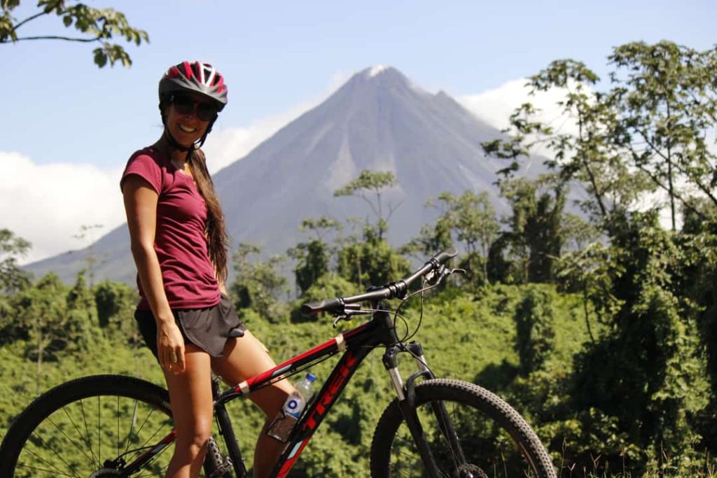 Lachende vrouw met fietshelm poseert naast haar mountainbike met de kegelvormige Arenal vulkaan op de achtergrond