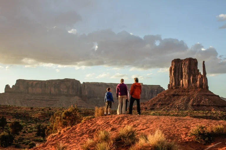 Gezin van drie personen staat op een rotsige heuvel en kijkt uit over de rode rotsformaties van Monument Valley in warm avondlicht