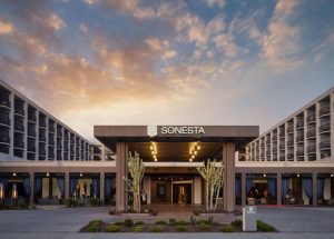 Hotelgebouw met een overdekte entree geflankeerd door hoge cactussen, verlicht bij zonsondergang met een kleurrijke lucht