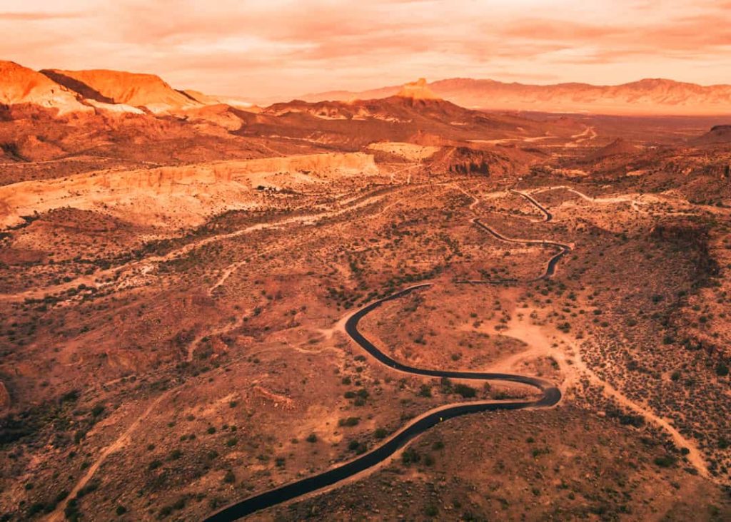 Luchtfoto bij zonsondergang van een smalle asfaltweg die kronkelt door een rood en okerkleurig woestijnlandschap met heuvels in Arizona