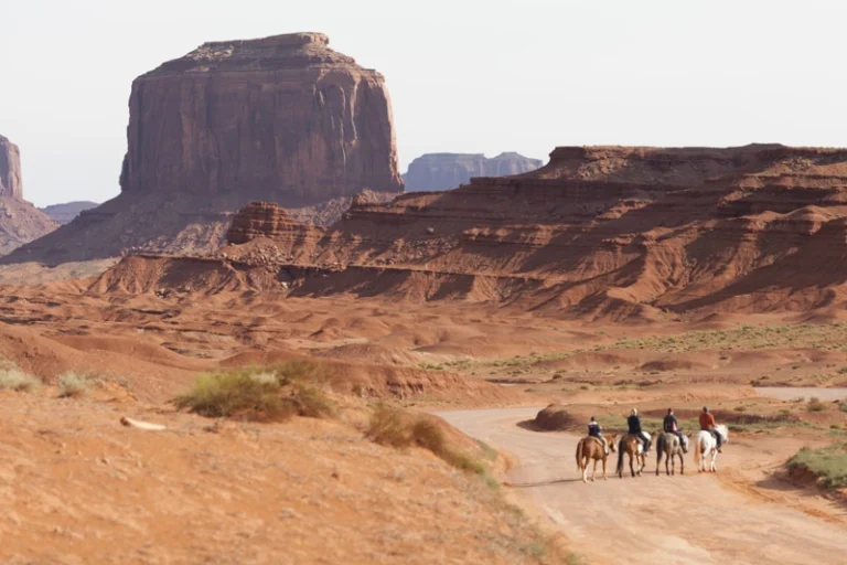 Vier ruiters te paard rijden over een zandweg met hoge rode rotsformaties van Monument Valley op de achtergrond