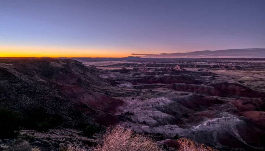 Wijds panorama van gelaagde heuvels van de Painted Desert onder een oranje gloeiende avondlucht met een opkomende maan