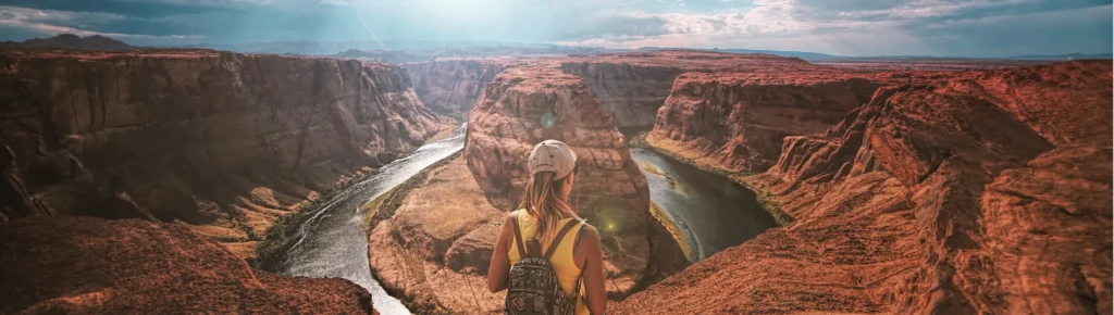 Vrouw met rugzak en pet staat op de rand van de canyon en kijkt uit over de kronkelende rivier bij Horseshoe Bend in Arizona