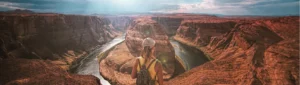Vrouw met rugzak en pet staat op de rand van de canyon en kijkt uit over de kronkelende rivier bij Horseshoe Bend in Arizona