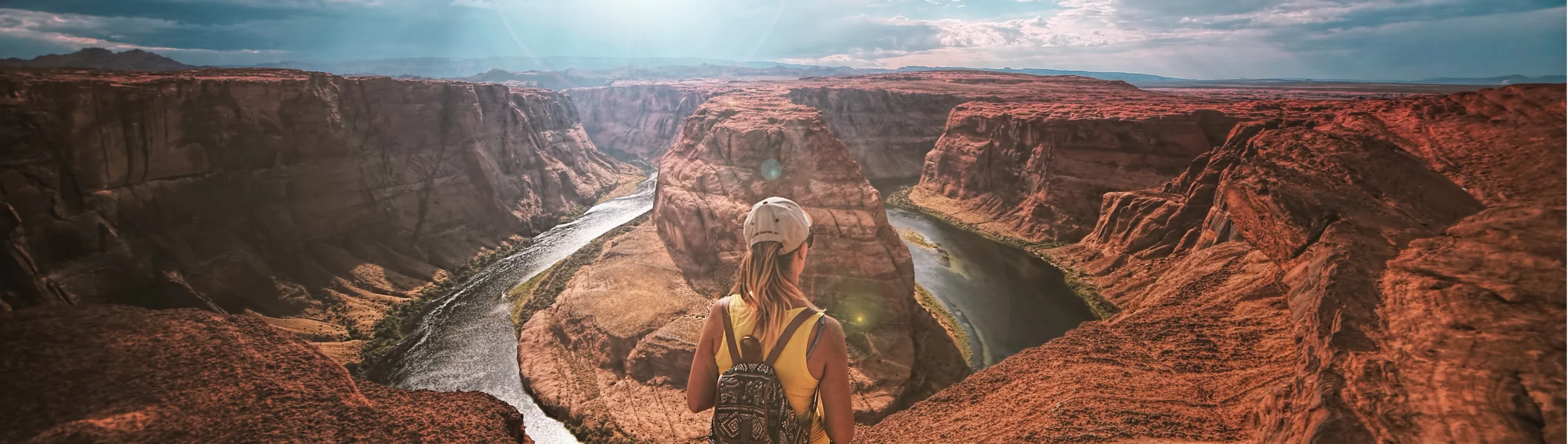 Vrouw met rugzak en pet staat op de rand van de canyon en kijkt uit over de kronkelende rivier bij Horseshoe Bend in Arizona