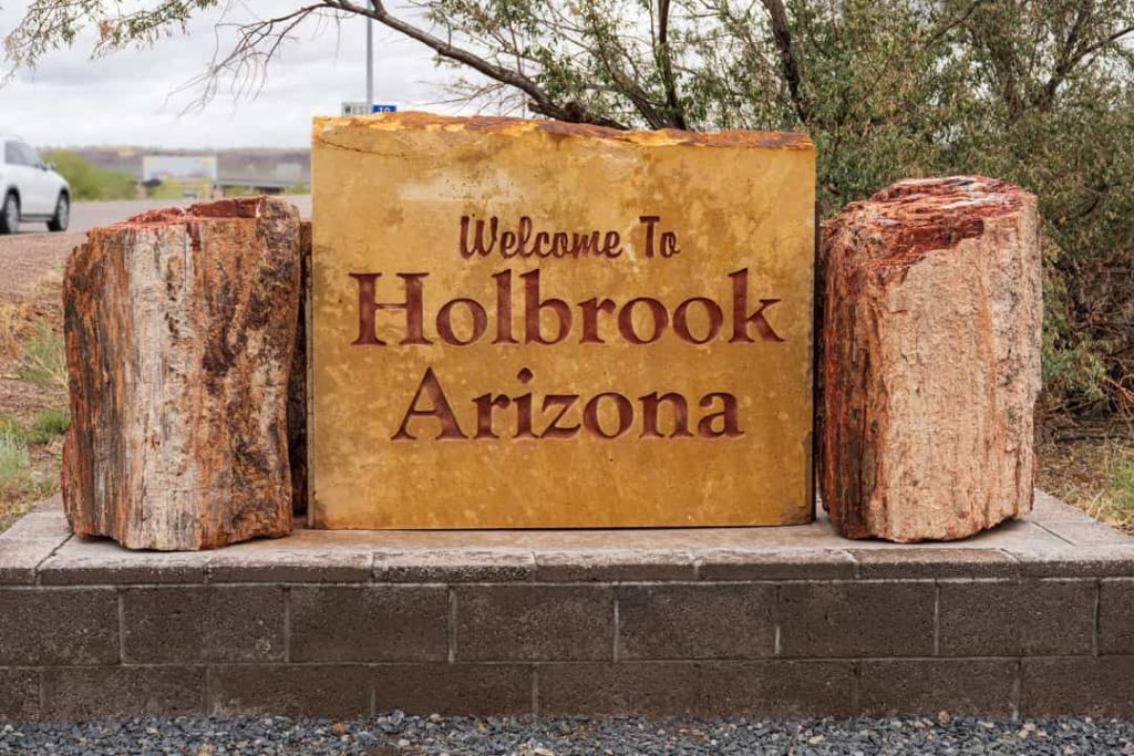 Stenen welkomstbord met de tekst Welcome to Holbrook Arizona tussen twee grote brokken versteend hout langs de weg
