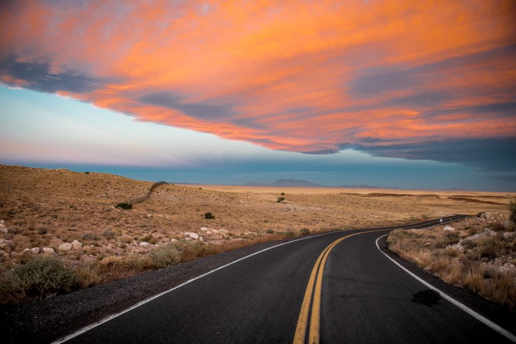 Bochtige tweebaansweg door een droog grasland met struiken, onder een dramatische oranje en blauwe avondlucht in Arizona