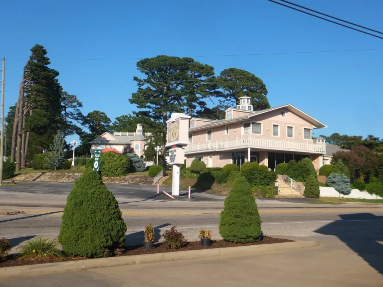 Een roze hotelgebouw met witte veranda's en trappen, omringd door struiken en hoge naaldbomen onder een strakblauwe hemel in Arkansas