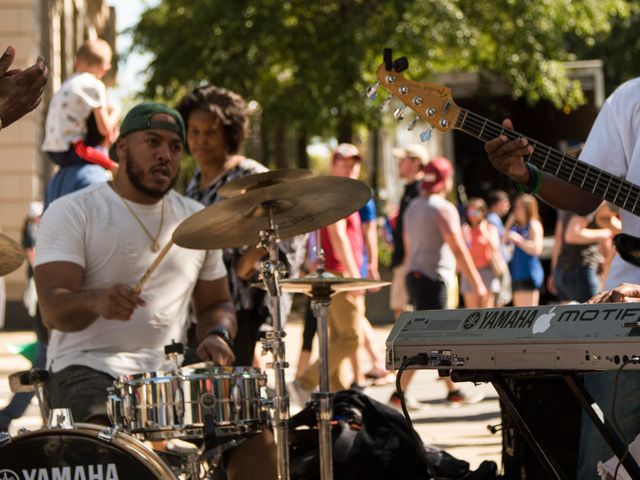 Een drummer in wit shirt speelt op een drumstel tijdens een straatoptreden in Arkansas, met publiek en bandleden op de achtergrond