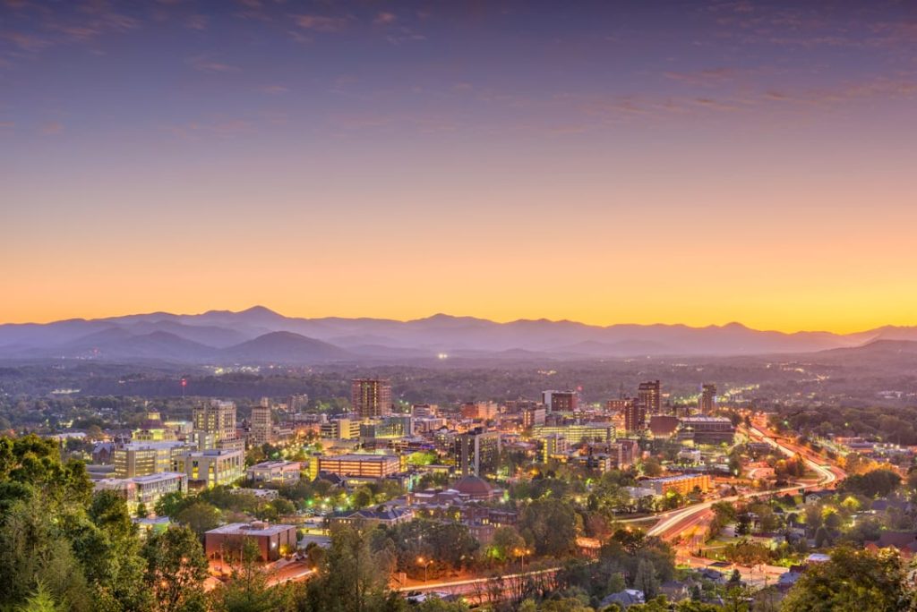 Downtown Asheville met gebouwen en verlichte straten voor een silhouet van de Blue Ridge Mountains bij zonsondergang