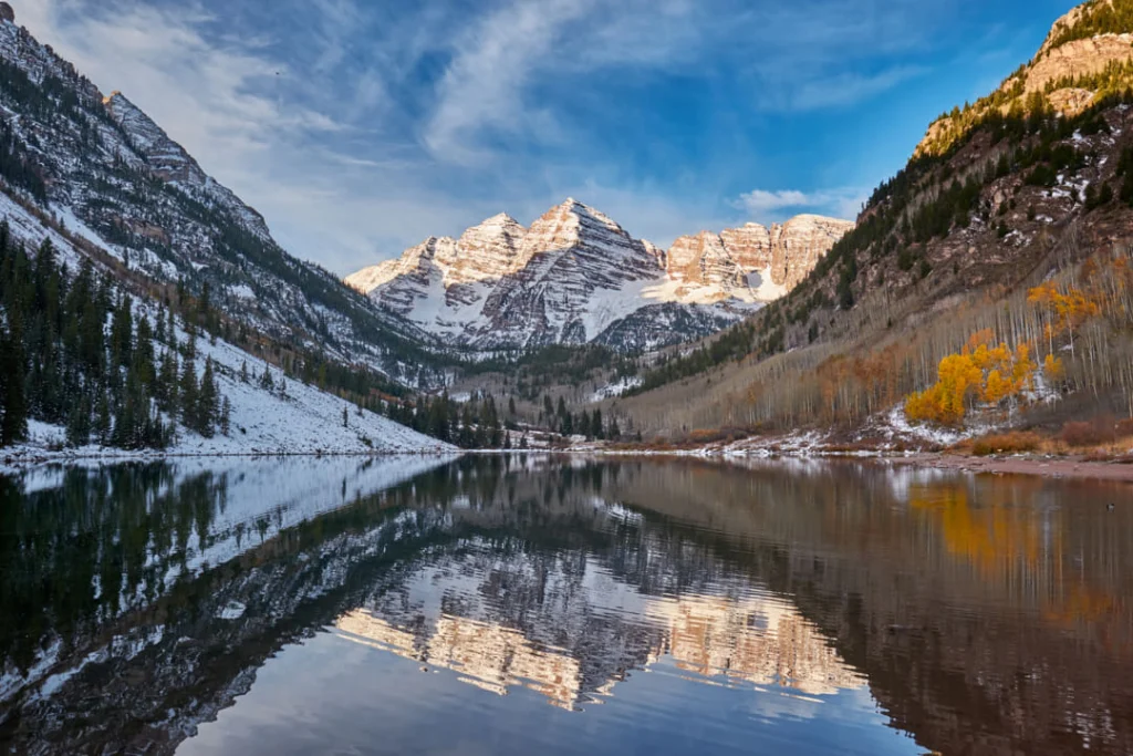 Besneeuwde toppen van de Maroon Bells weerspiegeld in een kalm meer, met goudgele espen aan de oever bij Aspen