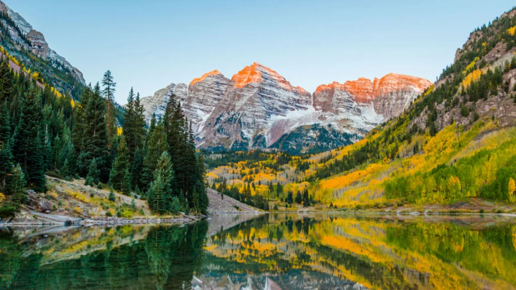 Twee markante bergtoppen van de Maroon Bells weerspiegeld in een rustig meer omgeven door herfstkleurig bos