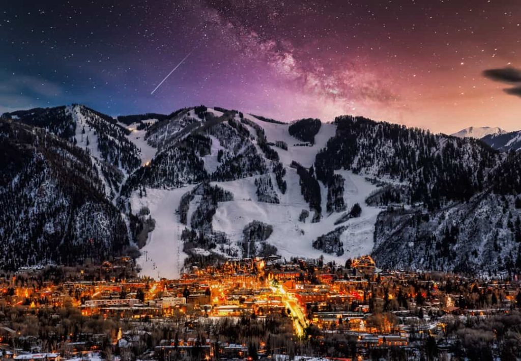 Verlicht dorp Aspen in Colorado gezien vanaf de bergen met besneeuwde skipistes en de melkweg zichtbaar aan de nachtelijke sterrenhemel