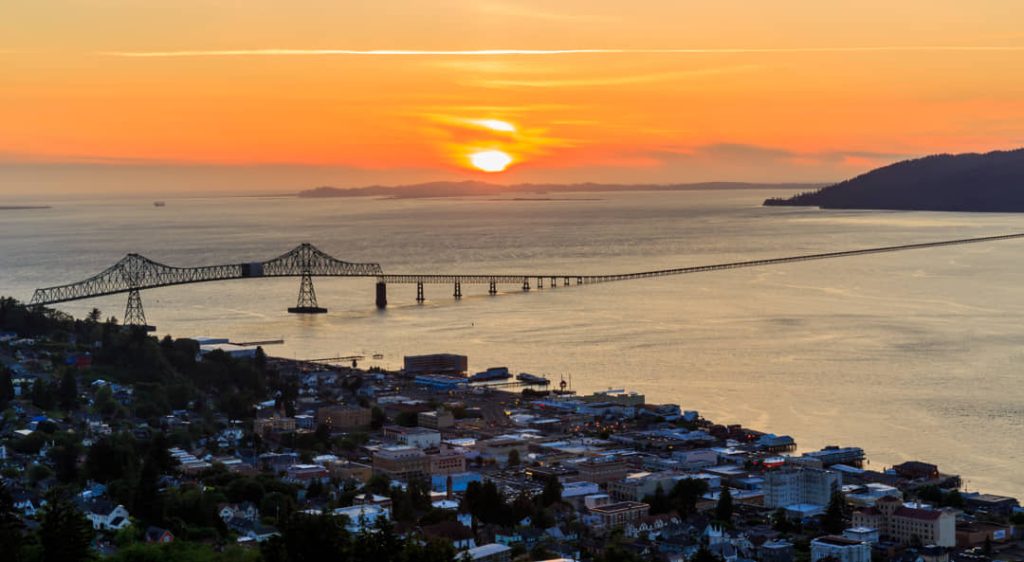 Luchtfoto vanaf een heuvel over de stad Astoria met bos, een kronkelende weg en de lange brug over de baai op de achtergrond