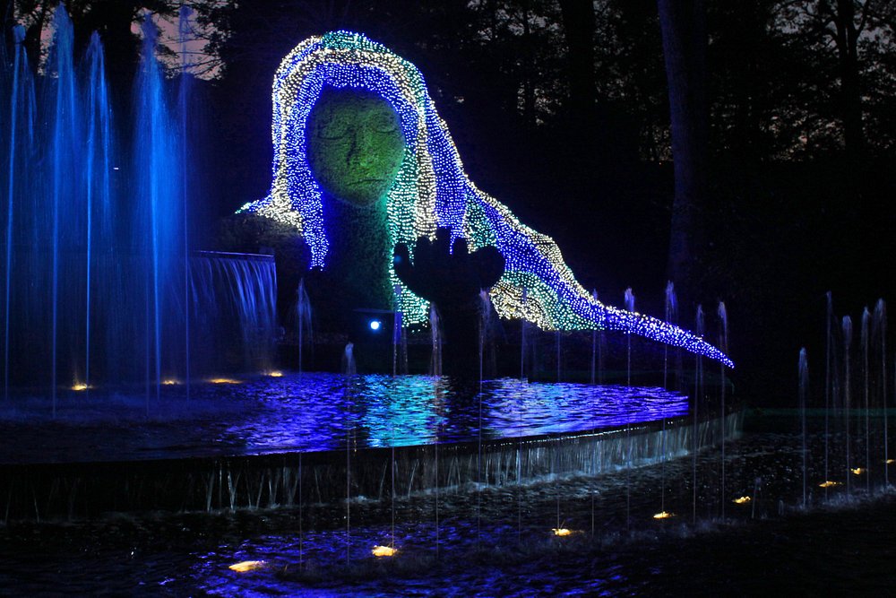 Grote lichtgevende sculptuur van een gesluierde figuur boven een vijver met fonteinen tijdens een avondlichtshow in een tuin
