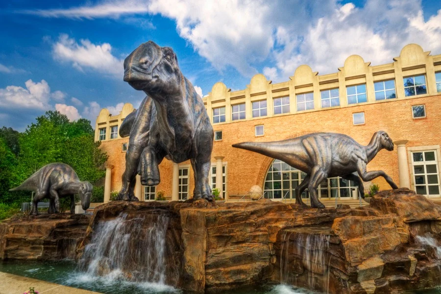 Drie grote dinosaurusbeelden staan op rotsen met watervallen voor een gebouw van het Fernbank museum in Atlanta