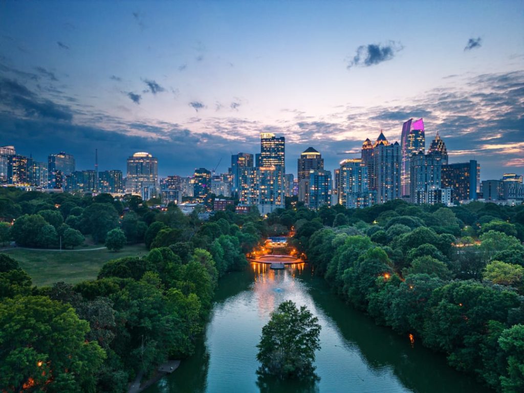 Verlichte wolkenkrabbers van Atlanta boven een groen park met een rivier en verlichte paviljoens op de voorgrond bij het invallen van de avond