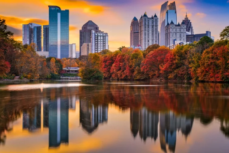 De wolkenkrabbers van Atlanta in Georgia weerspiegelen in een meer, omringd door bomen met oranje en rode herfstbladeren bij zonsondergang