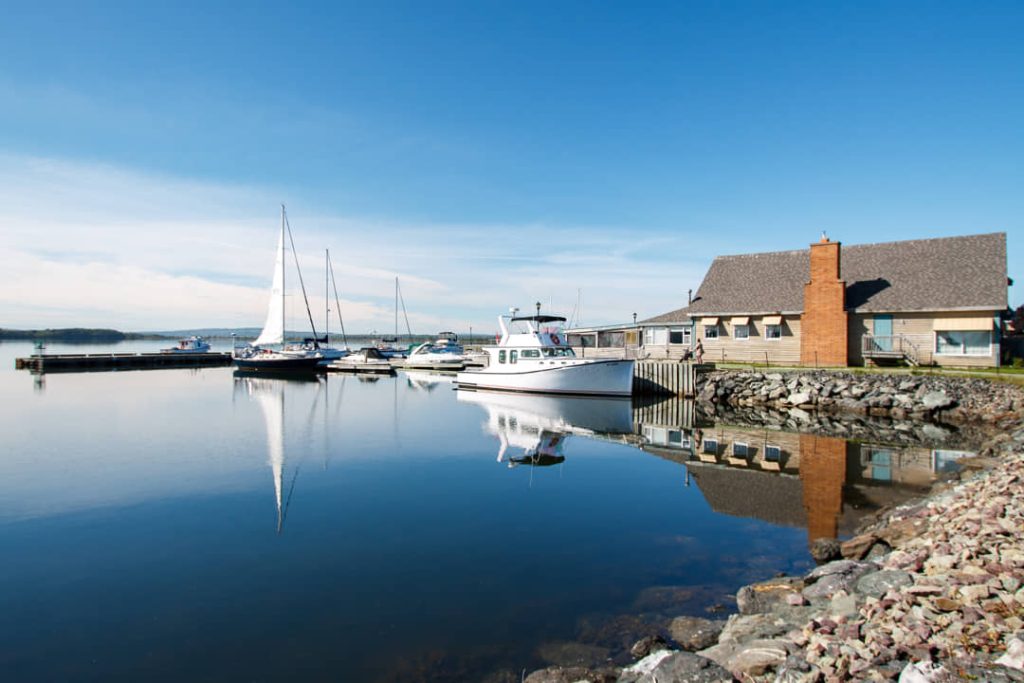 Zeilboten en een motorboot liggen afgemeerd bij een huisje met schoorsteen aan een rustig blauw water