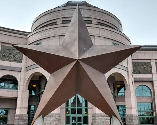Grote bronzen Texaanse ster op de voorgrond, met de ronde gevel van het Bullock Texas State History Museum erachter