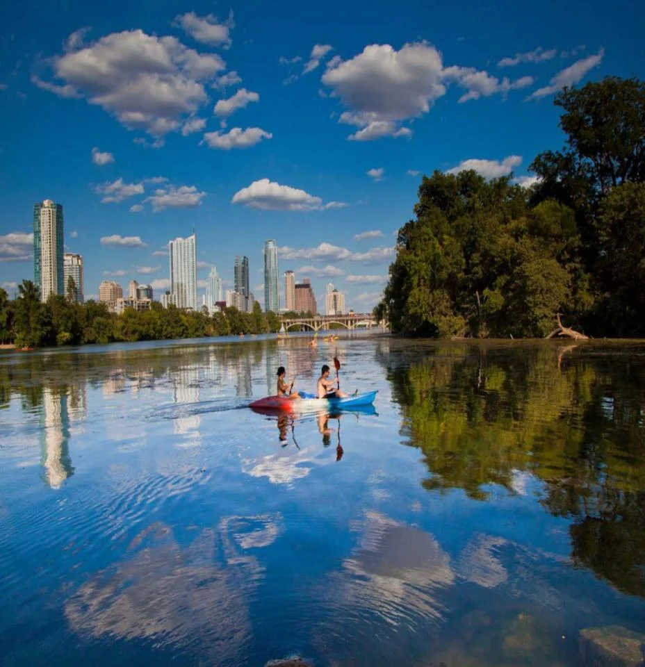 Twee mensen kajakken op het kalme water van Lady Bird Lake, met bomen en de skyline van Austin