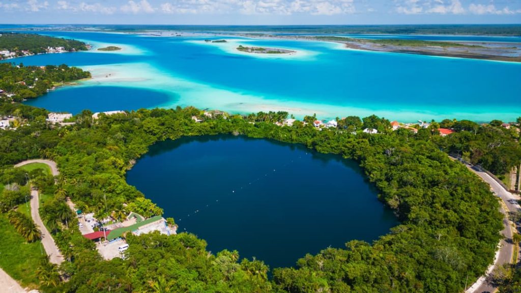 Luchtfoto van de donkerblauwe ronde cenote Azul omringd door groen naast de turquoise lagune van Bacalar