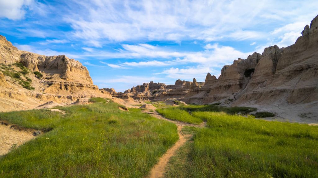 Grasrijk wandelpad tussen gele en grijze rotsformaties onder een deels bewolkte lucht in Badlands National Park