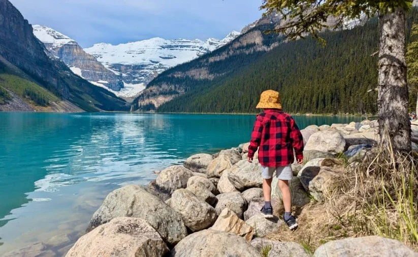 Een jongen in geruite rode jas en gele hoed staat op rotsen bij het turquoise water van Lake Louise, met besneeuwde bergen op de achtergrond in Banff National Park