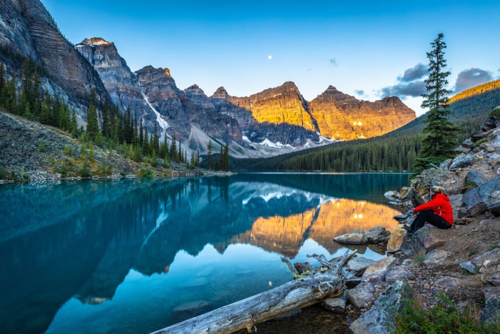 Wandelaar in rode jas zit op de rotsen bij Moraine Lake terwijl bergtoppen goud oplichten in de vroege ochtendzon