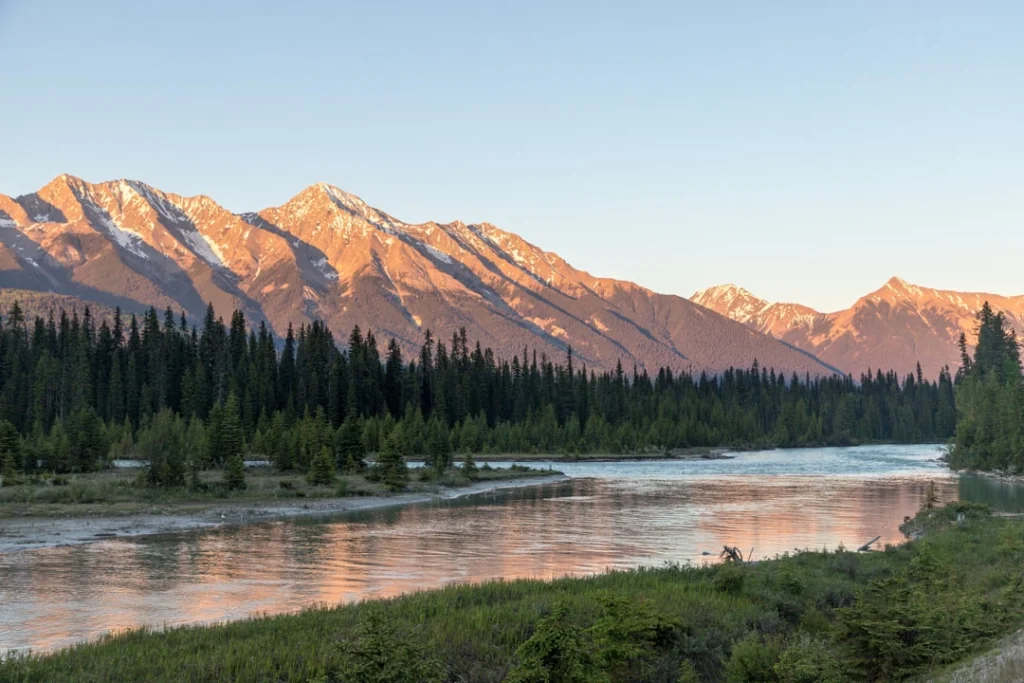 Rustige rivier tussen naaldbos, met rossig verlichte bergtoppen bij zonsopkomst in Banff National Park