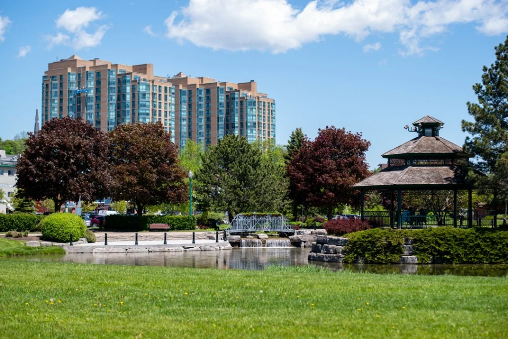Groen park met een vijver, bruggetje en houten paviljoen, met bomen en een hoge appartemententoren op de achtergrond in Barrie