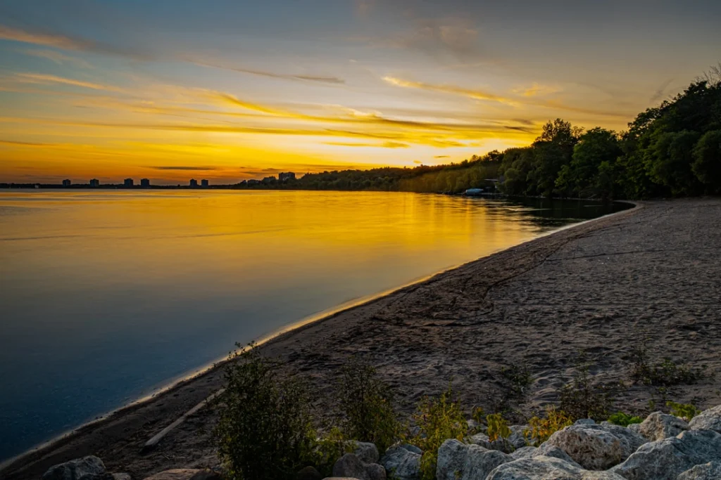 Zandstrand met stenen langs het meer in Barrie tijdens een gouden zonsondergang met bomen aan de waterlijn