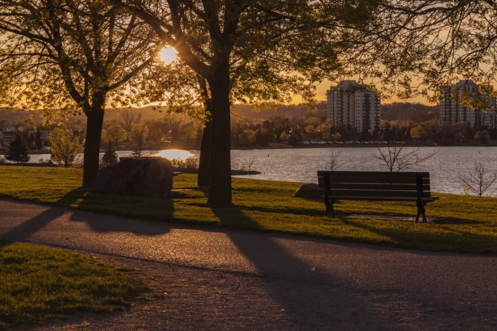 Bankje onder bomen in een park in Barrie met laagstaande zon en gebouwen aan de overkant van het water