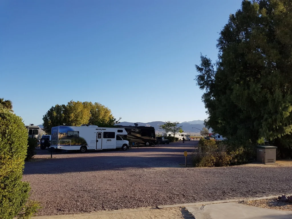 Verschillende campers staan geparkeerd op een grindterrein van een camping bij Barstow Calico met bomen en bergen op de achtergrond