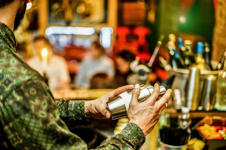 Een bartender schudt een cocktailshaker achter de bar, met flessen drank op de achtergrond