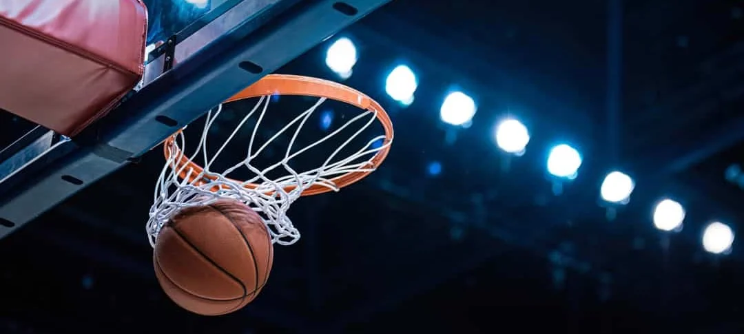 Close-up van een basketbal die door de basket gaat, met fel verlichte tribunelampen op de achtergrond