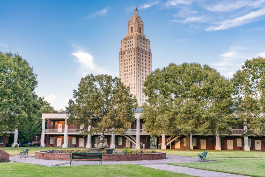 Rode bakstenen kazernegebouwen met zuilengangen rond een fontein met de hoge Capitol toren erachter in Baton Rouge